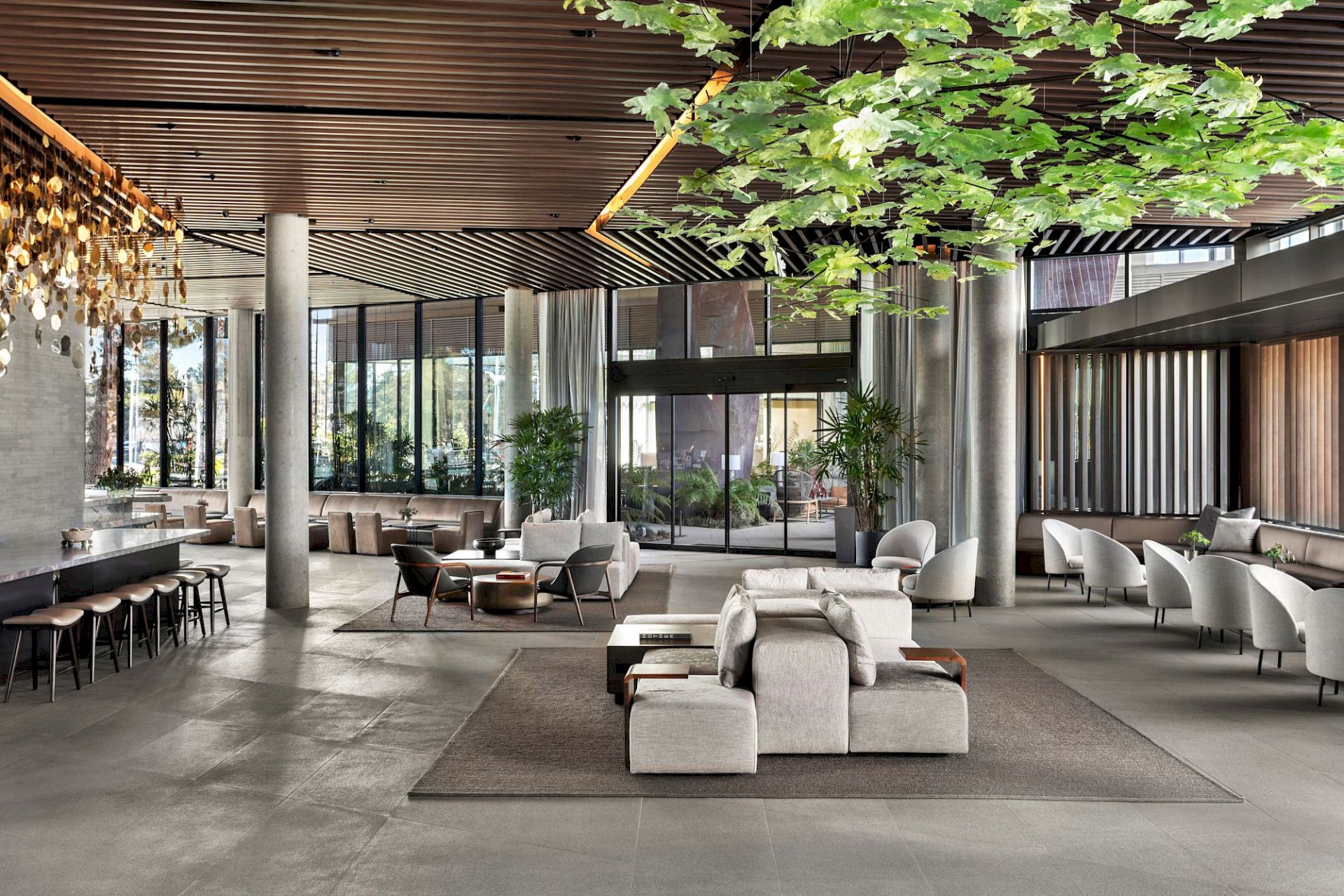 A modern lounge with sofas and plants, a hanging maple-like ceiling canopy, and tall windows filling a bright atrium.