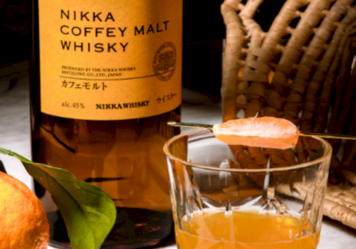 A bottle of Nikka Coffey Malt Whisky beside a glass of amber whiskey, with an orange and a wicker chair in the background.