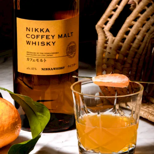 A bottle of Nikka Coffey Malt Whisky beside a glass of amber whiskey, with an orange and a wicker chair in the background.