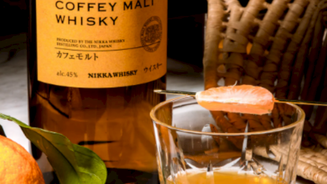 A bottle of Nikka Coffey Malt Whisky beside a glass of amber whiskey, with an orange and a wicker chair in the background.