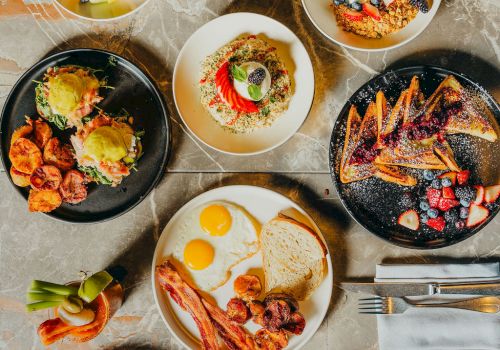 A brunch spread with sunny-side eggs, toast, roasted potatoes, and grilled meats, plus fruit, salads, and pancakes on plates and bowls.