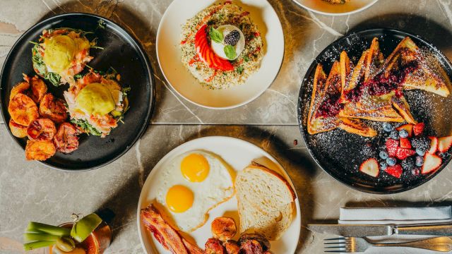 A brunch spread with sunny-side eggs, toast, roasted potatoes, and grilled meats, plus fruit, salads, and pancakes on plates and bowls.