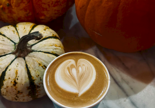 A latte with leaf-heart foam art beside autumn gourds and a carved pumpkin on a marble table.