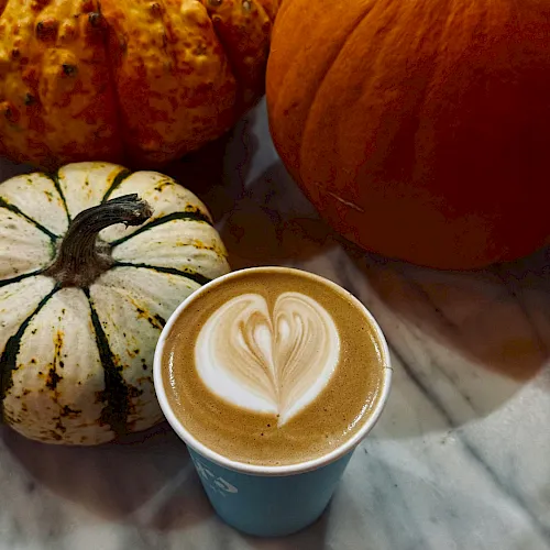 A latte with leaf-heart foam art beside autumn gourds and a carved pumpkin on a marble table.
