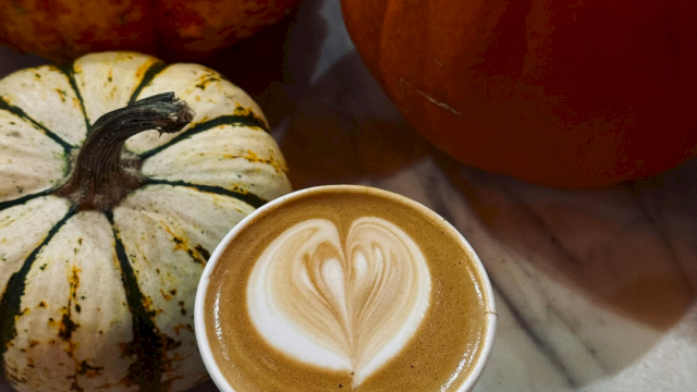 A latte with leaf-heart foam art beside autumn gourds and a carved pumpkin on a marble table.