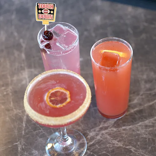 Three colorful cocktails: a pink drink, an orange drink, and a pink drink topped with a &ldquo;Touch Down&rdquo; flag.