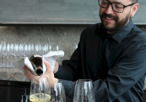 A man pours wine from a bottle into glasses at a tasting table during a presentation.