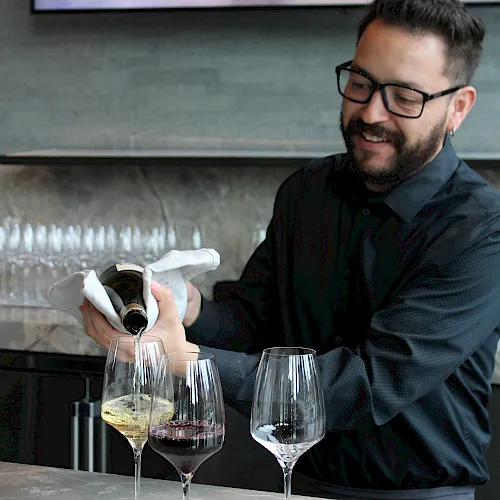 A man pours wine from a bottle into glasses at a tasting table during a presentation.