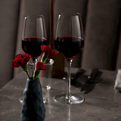 Two wine glasses filled with red wine, a blue vase with red flowers, and silver cutlery on a table in warm light.