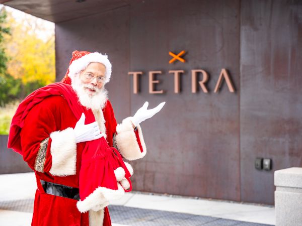 Santa Claus in a red coat stands beside a stone wall reading &ldquo;TETRA&rdquo; with an orange X over it.