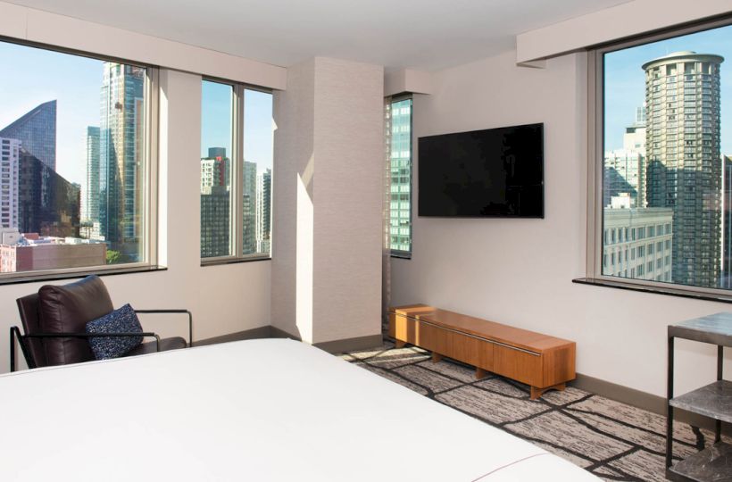 A modern hotel room with large windows overlooking a cityscape, featuring a bed, chair, wall-mounted TV, and minimalist decor.