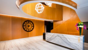 A modern interior with a marble front desk, wooden accents, a large wall clock, decorative lighting, and a flower arrangement.