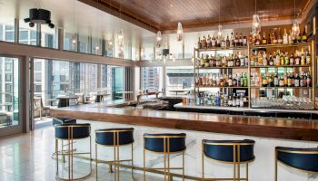 This image shows a modern bar with high chairs, a stocked back bar, and large windows offering a city view.
