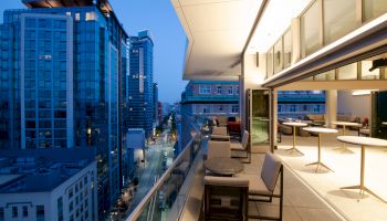 A modern city rooftop patio with tables and chairs overlooks a street lined with high-rise buildings at dusk.
