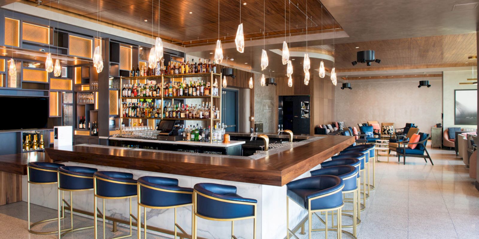 The image shows a modern bar with a counter, blue stools, numerous bottles on shelves, and pendant lights hanging from the ceiling.
