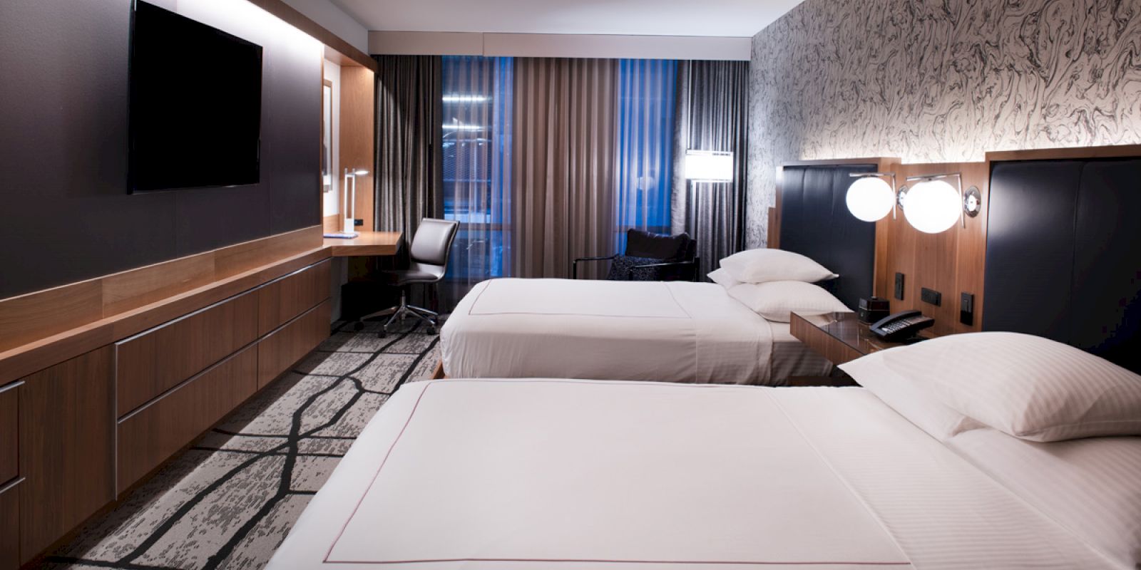 The image shows a modern hotel room with two beds, a flat-screen TV, desk, and ambient lighting.
