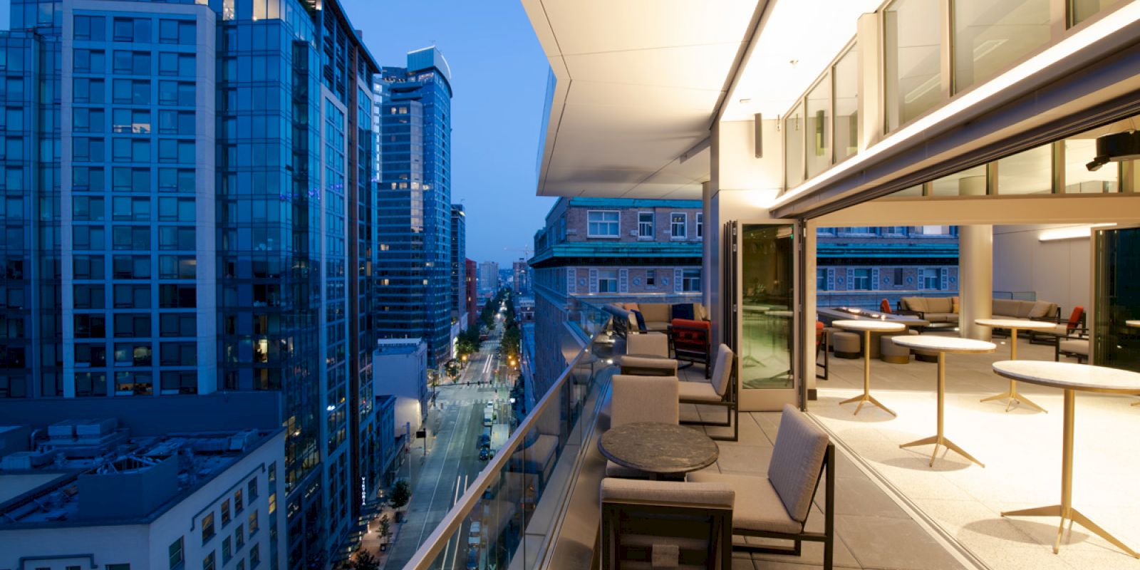 A modern outdoor terrace with seating overlooks a cityscape of high-rise buildings during evening.