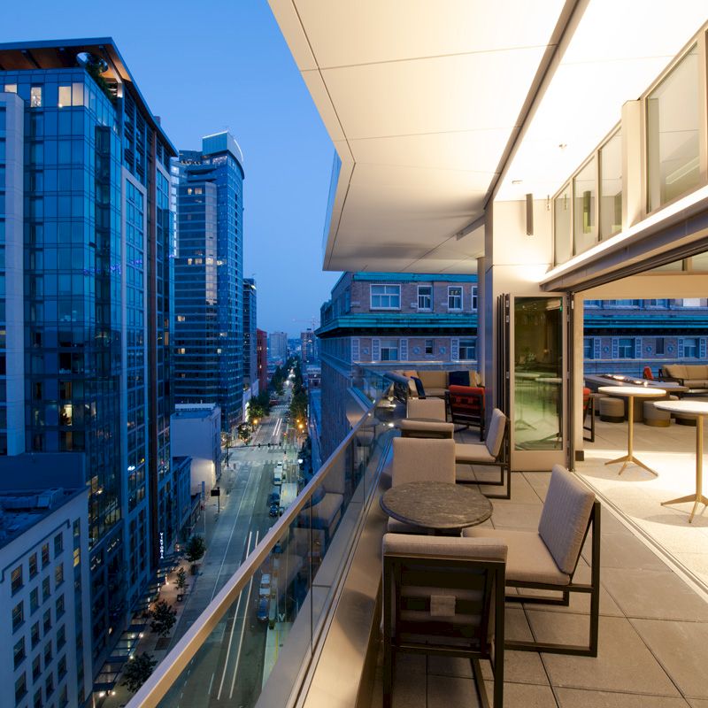 The image shows an urban rooftop view at dusk, featuring modern buildings, a furnished terrace, and a city street below.