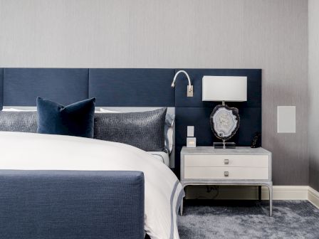 A modern bedroom with a blue headboard, white and blue bedding, a white bedside table, lamp, and wall-mounted reading light.