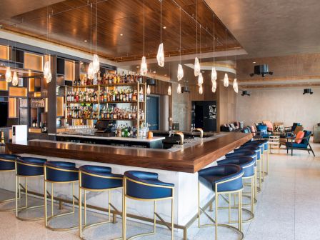A modern bar with a large selection of bottles, stylish pendant lights, blue bar stools, and a cozy seating area in the background.