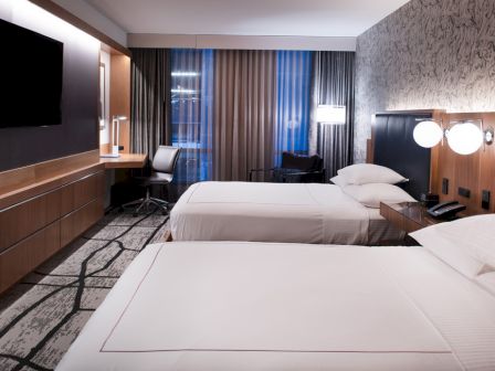 A hotel room with two beds, a desk, a TV, and modern decor, featuring patterned wallpaper and floor-to-ceiling curtains.