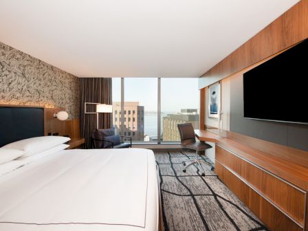 A modern hotel room with a large bed, desk, chair, flat-screen TV, and a window view of buildings and water in the distance.