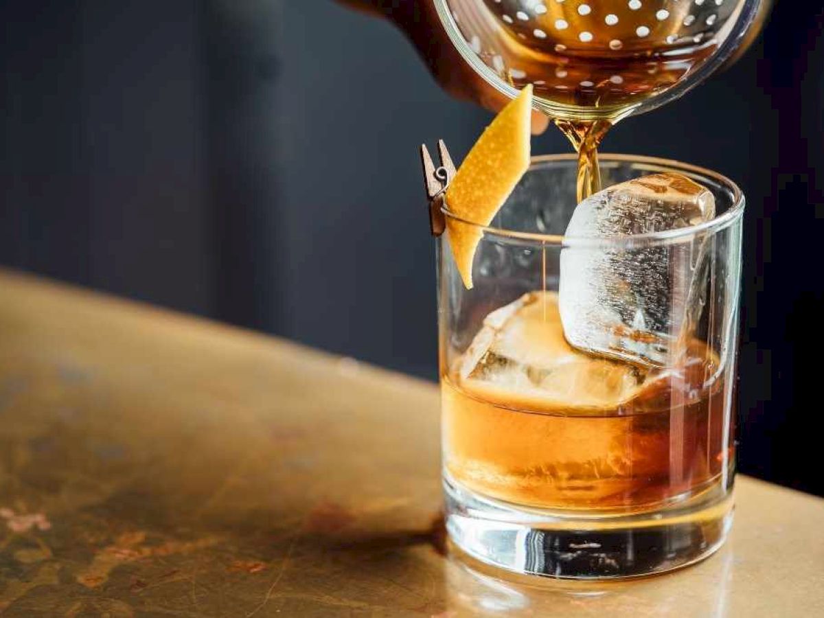 A cocktail with ice and a citrus garnish is being poured into a glass on a bar counter.
