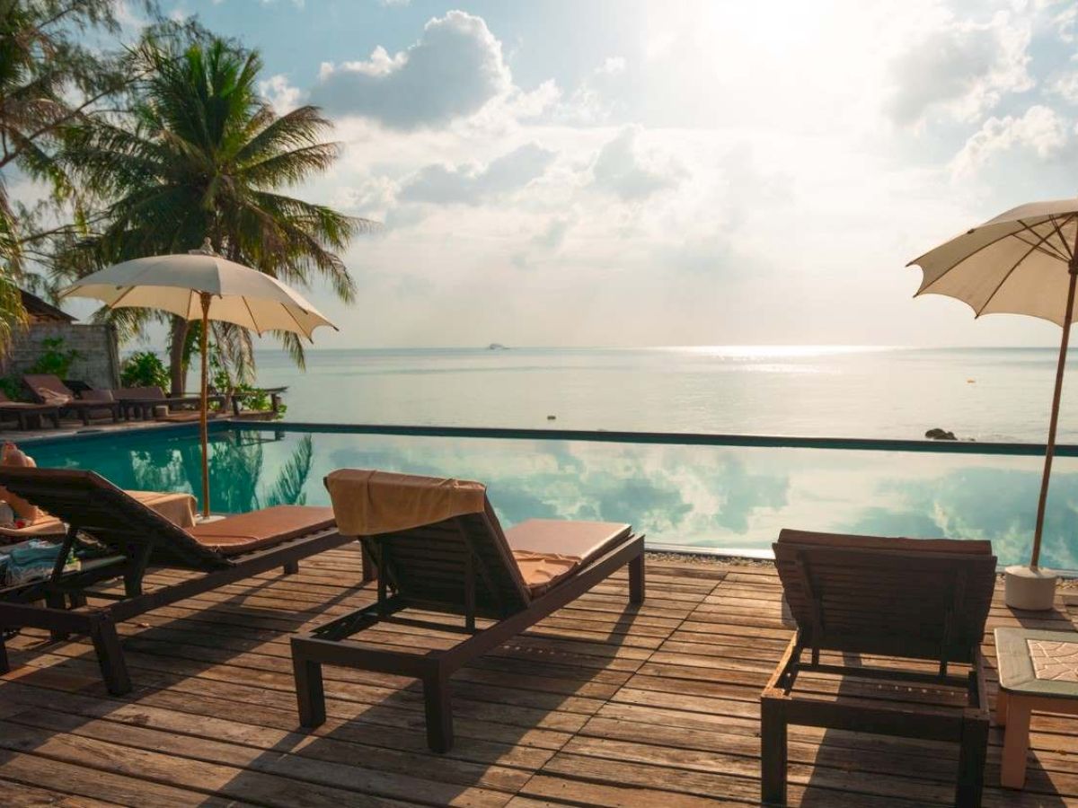 A serene tropical scene with lounge chairs, umbrellas, a pool, and views of the ocean under a cloudy sky, surrounded by palm trees.
