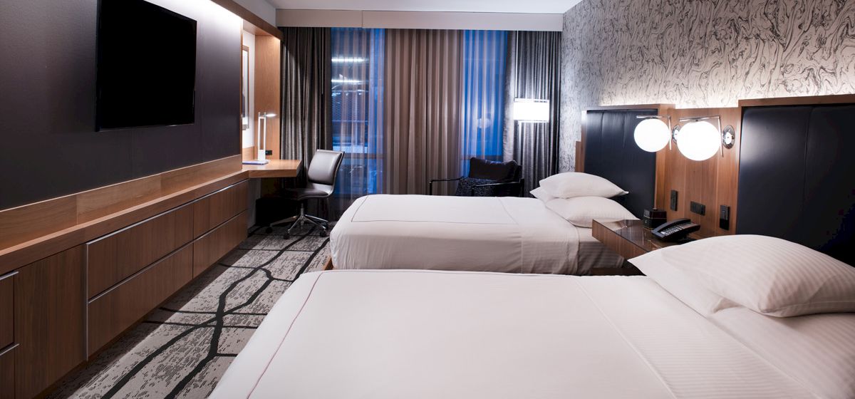 A modern hotel room with two beds, a wall-mounted TV, a desk, and lamps. The decor features wood accents and patterned walls.