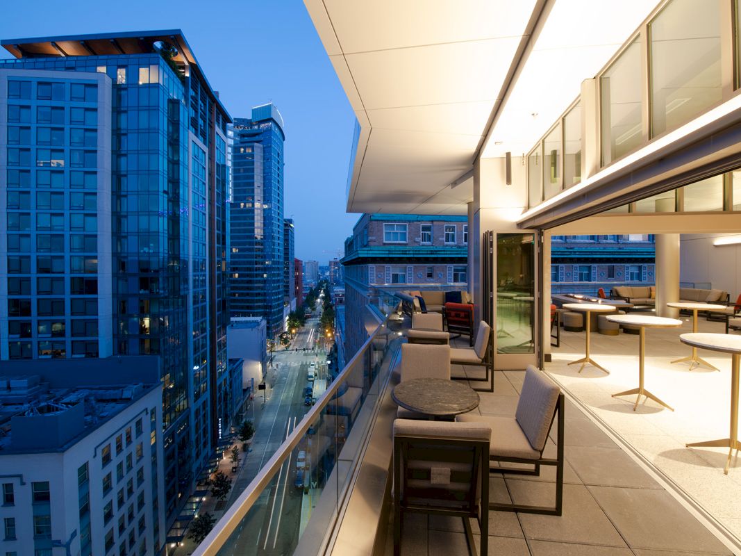 A modern cityscape at dusk, featuring tall buildings and a sleek balcony with tables and chairs, illuminated by warm indoor lighting.