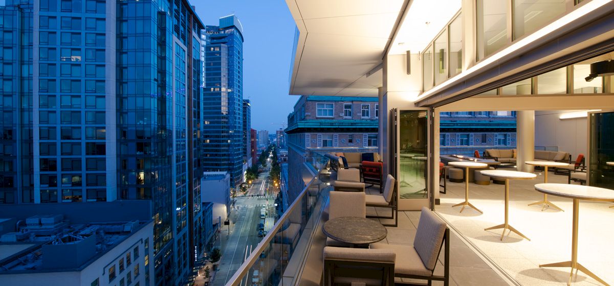 A modern cityscape at dusk, featuring tall buildings and a sleek balcony with tables and chairs, illuminated by warm indoor lighting.
