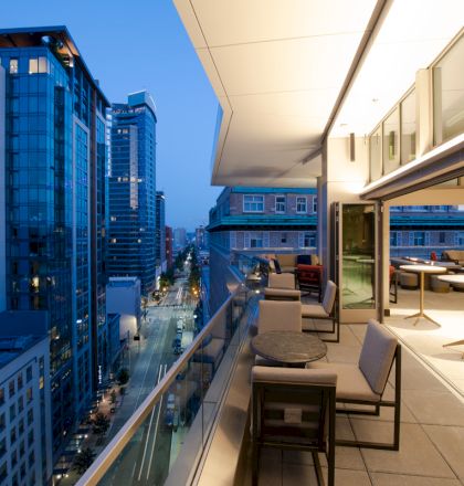 The image shows a modern outdoor patio on a high-rise building overlooking a cityscape at dusk, with chairs and tables along the balcony.
