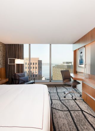 A modern hotel room with a large bed, wall-mounted TV, desk, and chair. The window offers a city and water view.