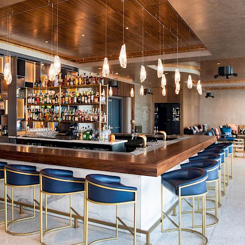 The image shows a modern bar with a stocked liquor shelf, stylish seating, pendant lights, and a spacious layout with additional seating area.