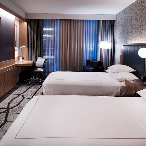 A modern hotel room with two neatly made beds, a wall-mounted TV, a desk with a chair, and patterned carpet and wallpaper.