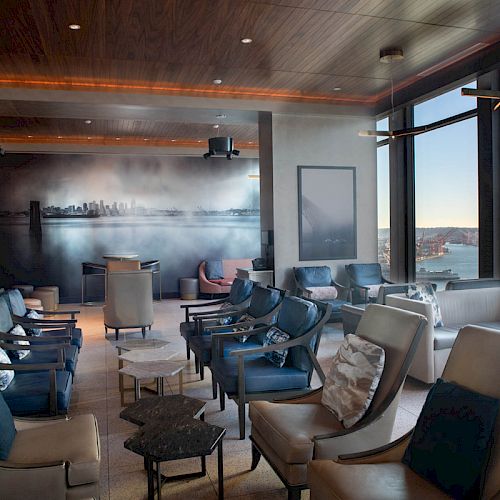 The image shows a modern lounge area with comfortable seating, a city mural on the wall, and large windows offering a view of a waterfront.