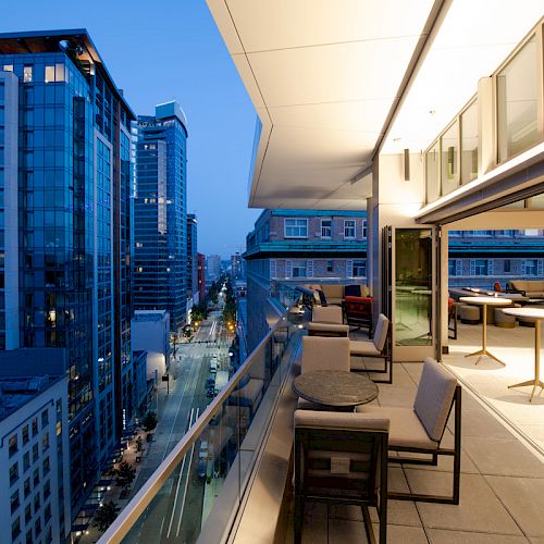A modern city view from a balcony at dusk, featuring high-rise buildings and a stylish open-air seating area with tables and chairs.
