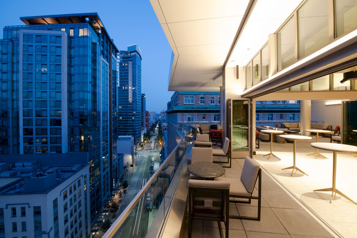A modern urban view from a balcony at dusk, featuring high-rise buildings and a stylish open-air seating area with tables and chairs.
