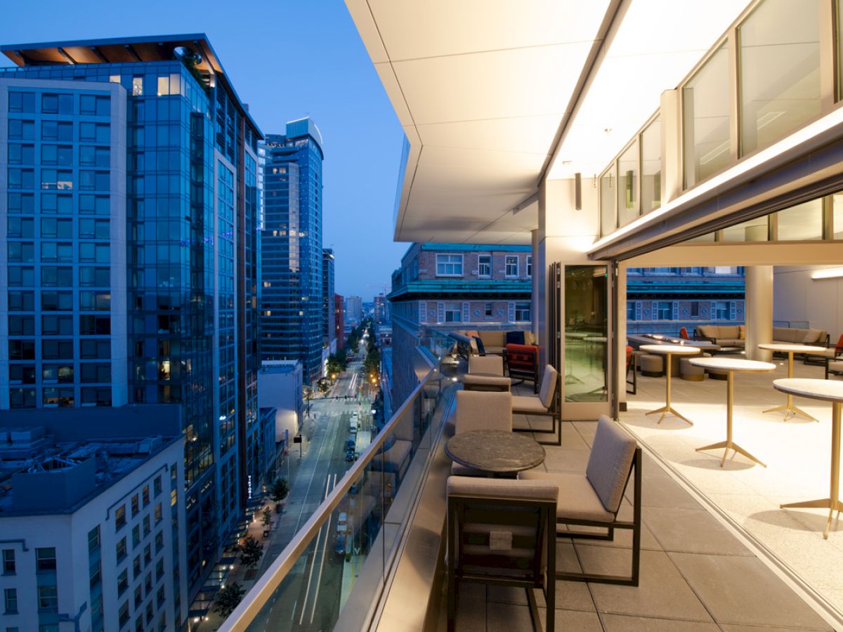 A modern urban view from a balcony at dusk, featuring high-rise buildings and a stylish open-air seating area with tables and chairs.