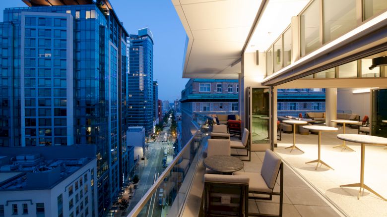A modern urban view from a balcony at dusk, featuring high-rise buildings and a stylish open-air seating area with tables and chairs.