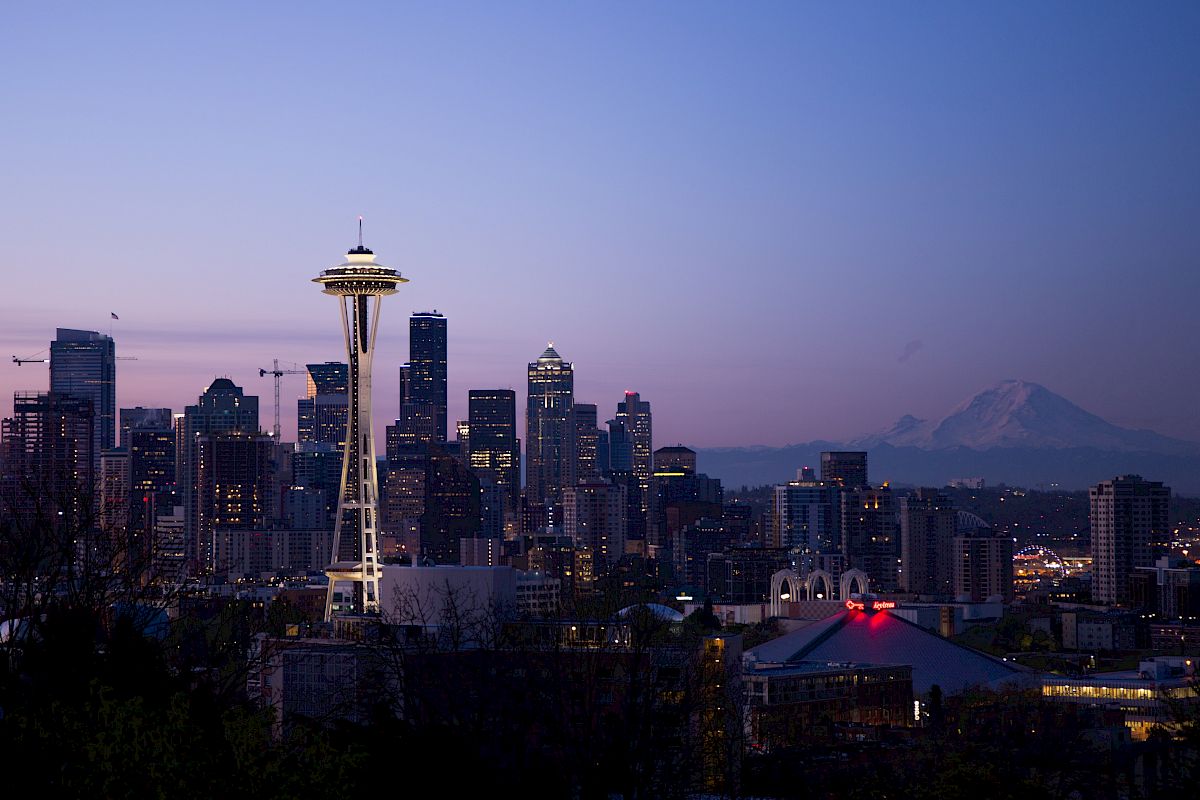 Seattle skyline at dusk with the Space Needle prominently visible and Mount Rainier in the background against a purple-toned sky.