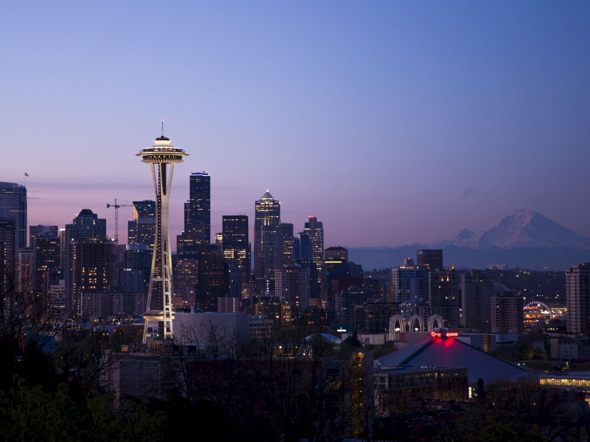 Seattle skyline at dusk with the Space Needle prominently visible and Mount Rainier in the background against a purple-toned sky.