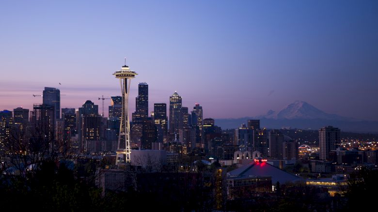Seattle skyline at dusk with the Space Needle prominently visible and Mount Rainier in the background against a purple-toned sky.