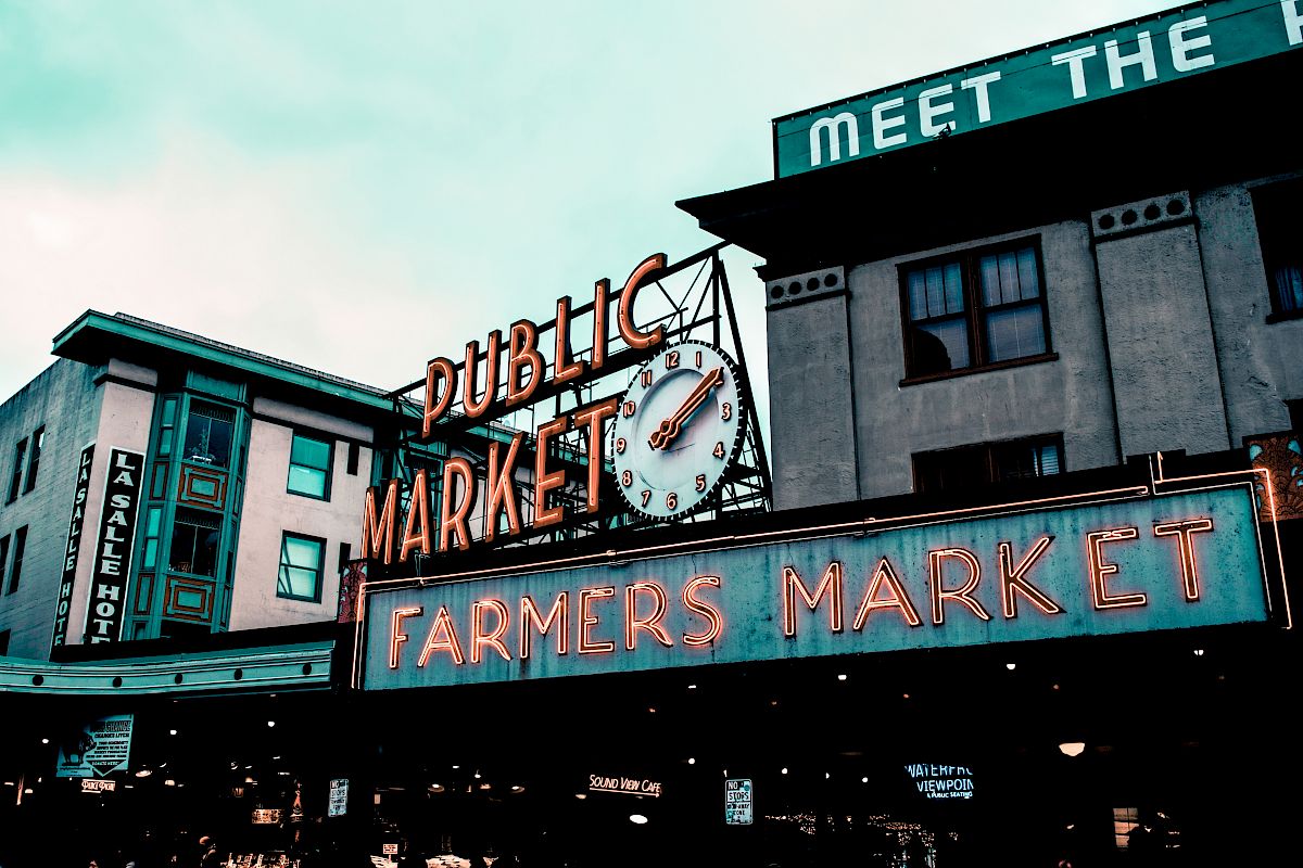 The image shows a public market with a notable clock and neon signs for a farmers market. A building with visible signage is in the background.