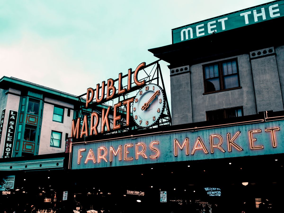 The image shows a public market with a notable clock and neon signs for a farmers market. A building with visible signage is in the background.