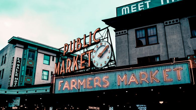 The image shows a public market with a notable clock and neon signs for a farmers market. A building with visible signage is in the background.