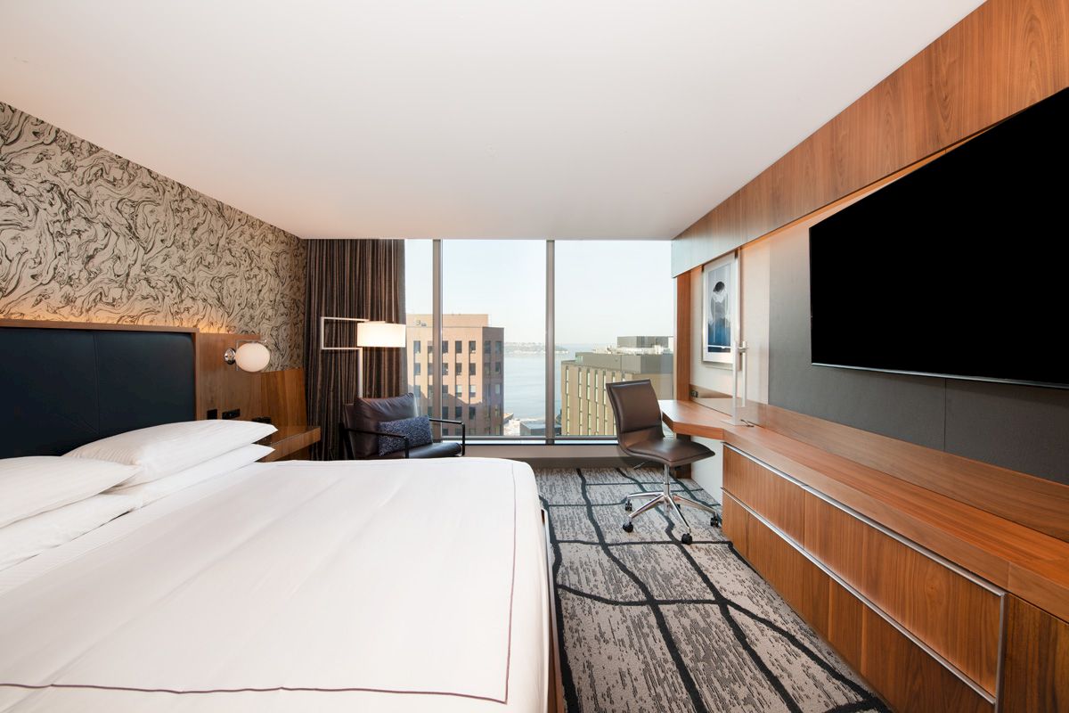 A modern hotel room features a large bed, wall-mounted TV, desk with chair, and a window with a cityscape view and striped carpet pattern.