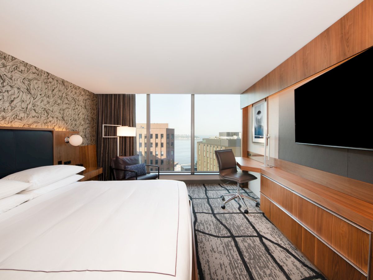 A modern hotel room features a large bed, wall-mounted TV, desk with chair, and a window with a cityscape view and striped carpet pattern.