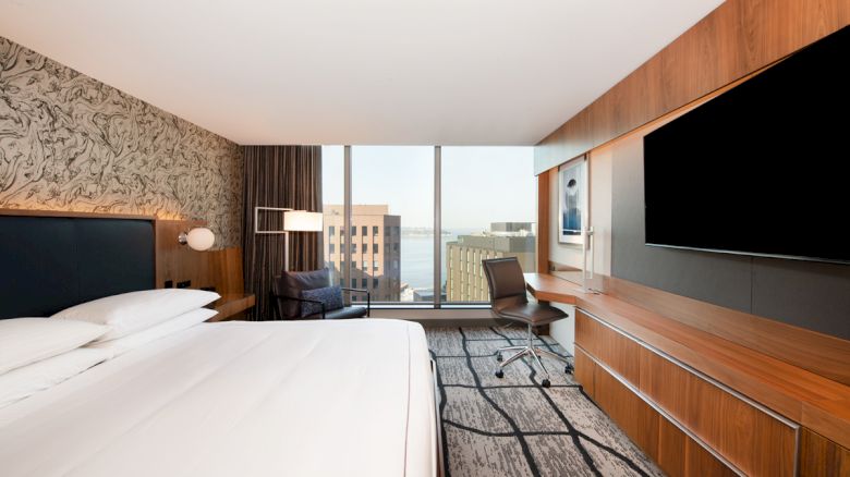 A modern hotel room features a large bed, wall-mounted TV, desk with chair, and a window with a cityscape view and striped carpet pattern.