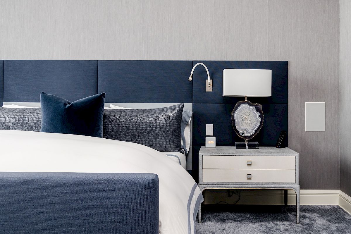 A modern bedroom with a blue and white color scheme, featuring a bed, pillows, nightstand, lamp, and wall-mounted reading light.